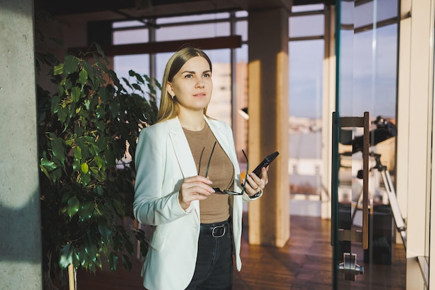 A successful young woman in glasses and a jacket is standing in the office and happily talking on the phone A young manager works in the office and manages the business remotely