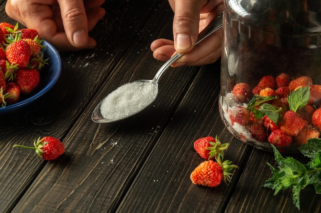 Sugar is added to jar of ripe strawberry and mint compote Hands of chef with spoon on kitchen table