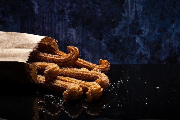 Sugared churros on black table with reflection. Churros in paper bag. Blue background.