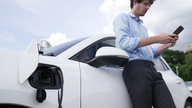 Suitclad businessman with progressive ambition leaning on his electric vehicle while standing on a charging station with a power cable plug and a renewable energypowered electric vehicle