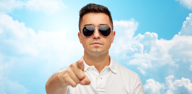 summer, accessories, style, gesture and people concept - face of middle aged latin man in white polo t-shirt and sunglasses pointing finger on you over blue sky and clouds background