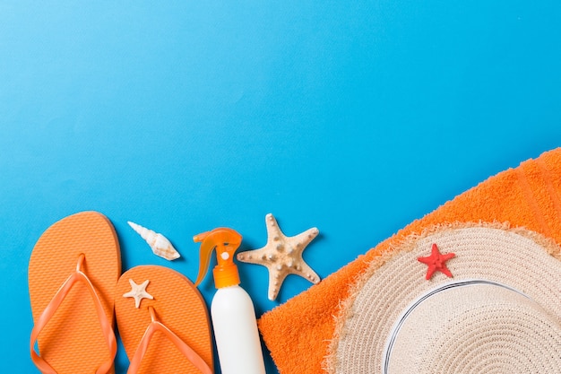 Summer beach flat lay accessories. Sunscreen bottle cream, straw hat, flip flops, towel and seashells on colored Background. Travel holiday concept with copy space.