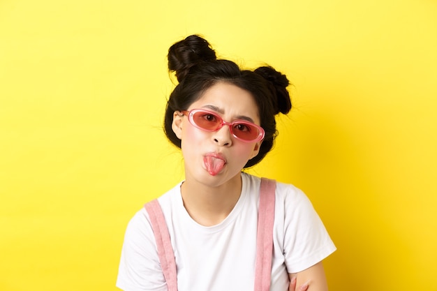 Summer and beauty concept. Childish asian girl in sunglasses showing tongue, being rude, standing on yellow.