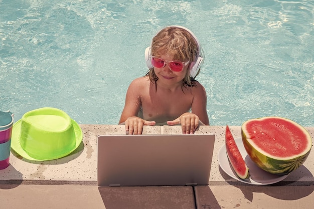 Summer business Child remote working on laptop in pool Little business man working online on laptop in summer swimming pool water Little businessman working on tropical beach