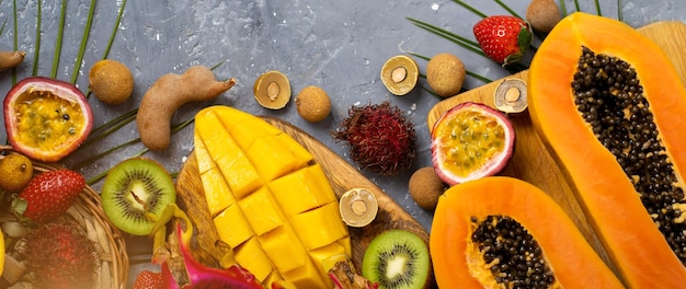 Summer composition of sliced in half papaya, dragon fruit, rambutan, tamarind, mango, passion fruit, kiwi, strawberry, leaves. Flat lay. Top view of tropical fruits on grey blue background.