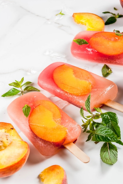 Summer desserts. Frozen drinks. Sweet fruit popsicles from frozen peach tea with mint. On a white marble table, with ingredients  peaches, mint, ice. 