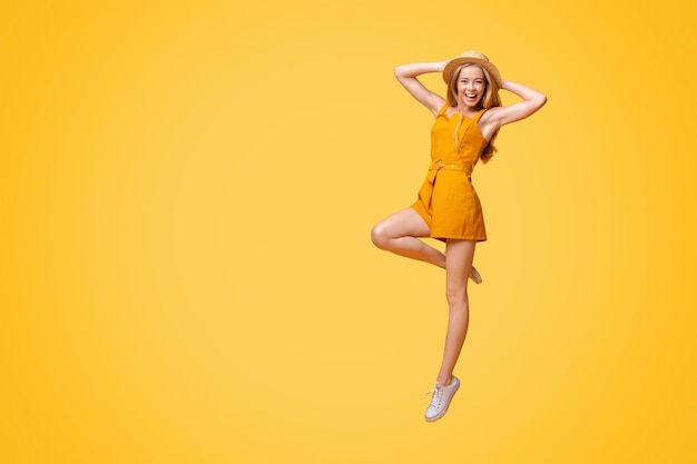 Summer fashion joyful teen girl in jumpsuit and straw hat jumping on yellow studio background with