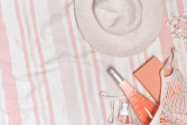 Summer flatlay with widebrimmed sun hat cosmetic product bottle rose wine book on striped beach towel as background Summer holiday vacation rest concept White pink pastel top view photo