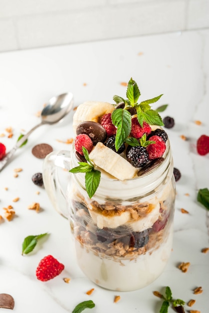 Summer fruit berry breakfast. Healthy banana split breakfast with cream cheese, raspberries, blackberries, mint, white and pink chocolate. In mason jar, marble table. Copy space close view