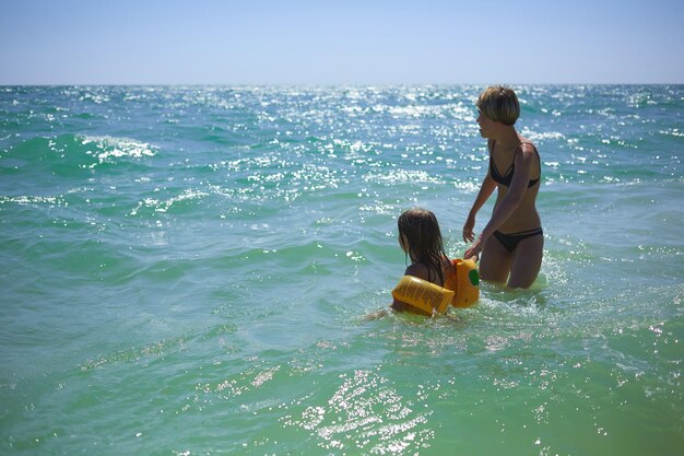 Summer happy family of six years blonde child playing and jumping water waves embracing woman mother in sea shore beach