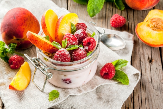 Summer healthy breakfast. Overnight oatmeal in a jar, granola, nuts, fresh raspberries and peach, decorated with mint leaves. On the old rustic wooden table. Copy space