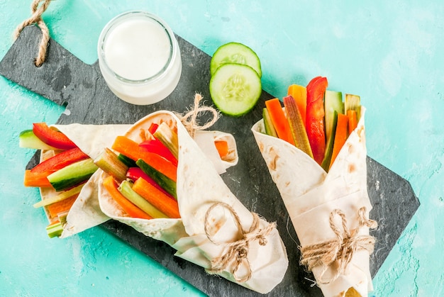 Summer healthy snack Mexican style tortilla sandwich wraps assorted colorful fresh vegetable sticks (celery rhubarb pepper cucumber and carrot) with yoghurt sauce dip light blue background