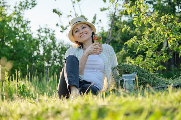 Summer holiday in garden, woman gardener sitting on freshly cut grass, resting female with fresh herbal natural berry drink