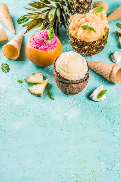 Summer holiday vacation concept, set various tropical ice cream sorbets, frozen juices in pineapple, grapefruit and coconut, light blue concrete 