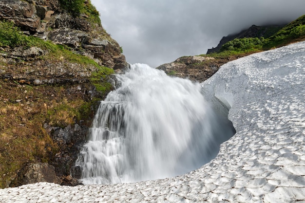 Summer landscape mountain waterfall falling into the snowfield