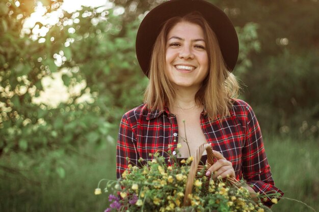 Summer lifestyle portrait of a beautiful young woman in a hat smiling and holding a basket with a bouquet of wild flowers The concept of happiness and love