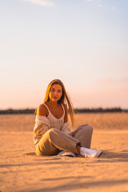 Summer lifestyle, a young blonde with straight hair, in a small wool sweater and corduroy pants sitting on the beach. Sitting in the serious sand