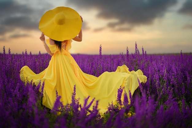 Summer mood. A woman in a luxurious yellow dress is standing in a purple flowering field with her back to the camera.