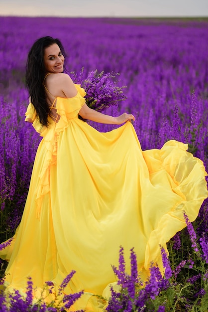 Summer mood. A woman in a luxurious yellow dress walks along a purple blooming field with her back to the camera.