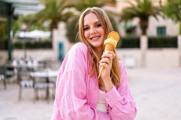 Summer outdoor portrait of pretty blonde woman eating tasty Italian gelato cone ice cream enjoy her vacation pink trendy outfit