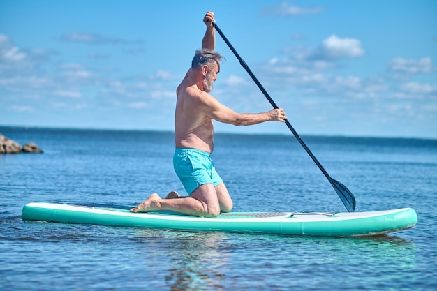 Summer pastime. Adult grey-haired bearded man kneeling sideways to camera paddling on board on water on sunny day