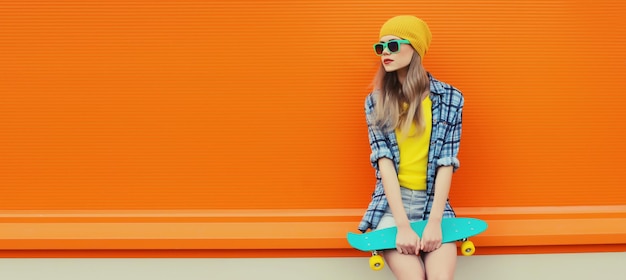 Photo summer portrait of stylish young woman with skateboard in colorful clothes on orange background