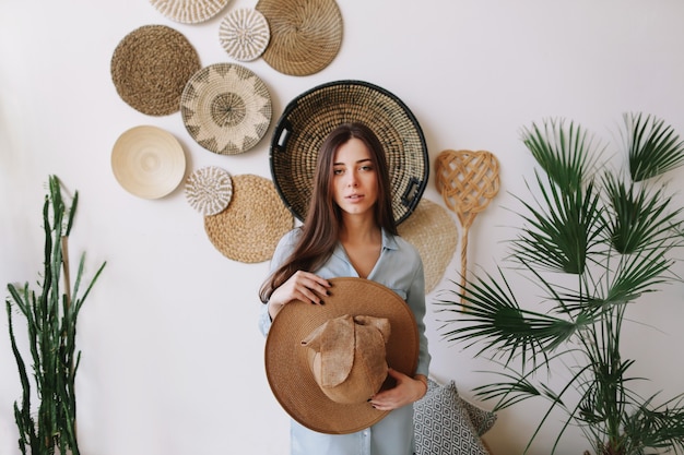 Summer portrait of a young pretty woman in a dress and holding a hat in exotic boho interior