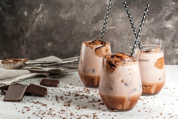 Summer refreshment drinks. Chilled iced chocolate cocoa. With scoop of chocolate ice cream, chocolate powder and ice. In glasses, with tubes for drinking. White concrete table. 