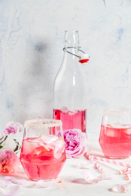 Summer refreshment drinks. Light pink rose cocktail, with rose wine, tea rose petals, lemon. On a white stone concrete table. With striped pink tubules, petals and rose flowers. Copy space