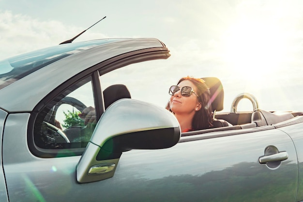 Summer road trip She travels to a gig with open roof Satisfied Woman sitting relaxed in the car against the sky with bright sun concept of traveling by car Summer holiday driving pleasure