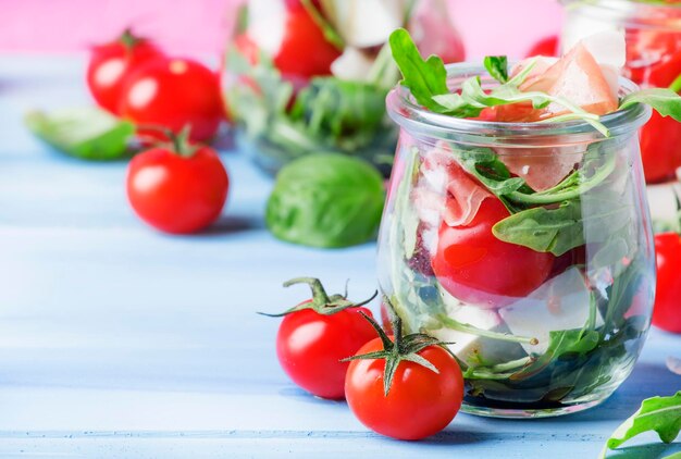 Summer salad with arugula soft cheese cherry tomatoes and prosciutto in glass jars festive background selective focus