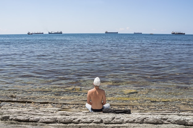 Summer sea landscape. A guy sits on a stone wild beach and looks into the distance. On the horizon is a clear blue sky and cargo ships.