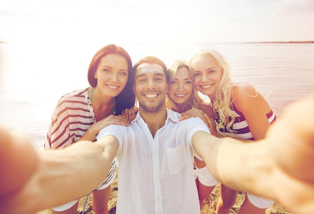 summer, sea, tourism, technology and people concept - group of smiling friends with camera on beach photographing and taking selfie