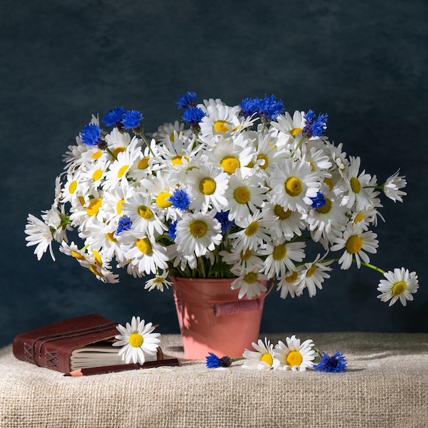 Summer still life A large bouquet of daisies and cornflowers in a pink bucket Nearby is a notebook and a pencil on a blue background