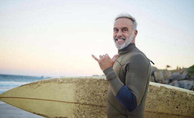 Summer surfing and portrait of old man at the beach in the morning Retired senior man with surfboard hand sign and smile on face ready to surf at sunrise Ocean hobby and water sport in Australia