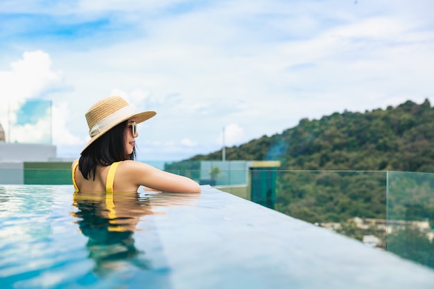 Summer travel vacation concept, Happy traveler asian woman with hat and bikini relax in luxury infinity pool hotel resort with sea beach background at day in Phuket, Thailand