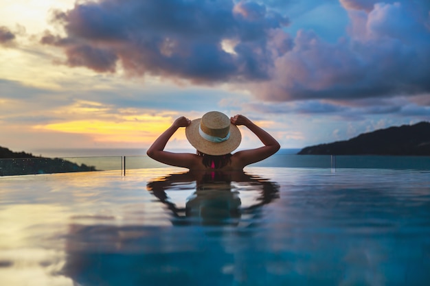 Summer travel vacation concept, Happy traveler asian woman with hat and bikini relax in luxury infinity pool hotel