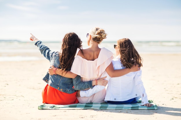 summer vacation, holidays, travel and people concept - group of young women sitting on beach blanket and pointing finger to something