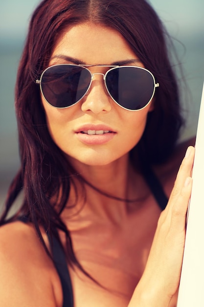 summer vacation, travel, surfing, water sport and people concept - young woman in sunglasses with surfboard on beach