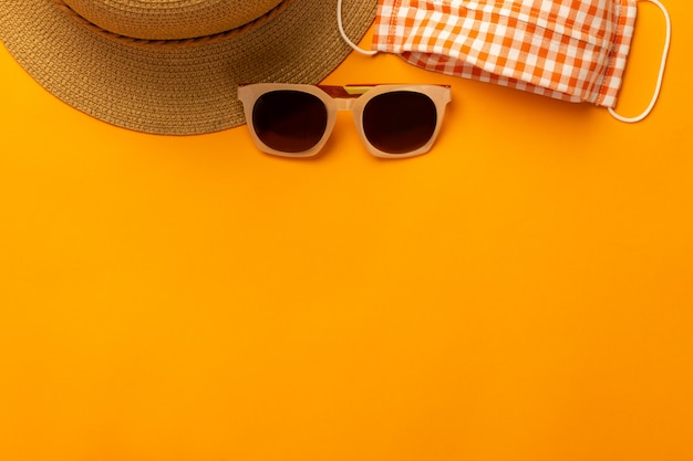 Summer wall with beach accessories - straw hat, sunglasses, mask to prevent covid-19 on vibrant orange wall top view with copy space