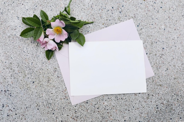 Summer wedding stationery mockup scene Horizontal blank greeting card invitation pink envelope and blooming dog rose flowers on textured terrazzo background Flat lay top view No people