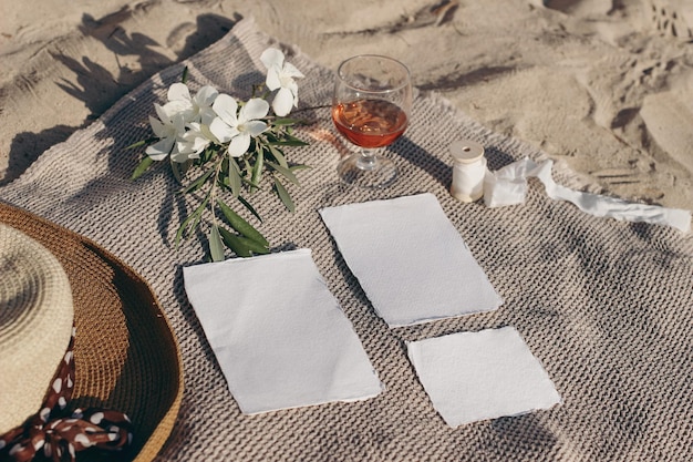 Summer wedding stationery mockups set Vacation still life Picnicon Mediterranean beach sand Straw hat white oleander flowers with olive tree branches rose wine glass on beige towel Top view