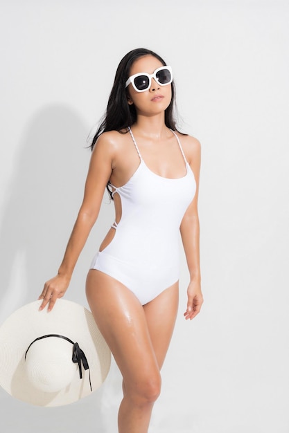 Summer woman studio portrait. Asian woman wearing white swimsuit dress in a standing position, holding a white white hat and sunglasses, in a summer fashion on isolated white background.