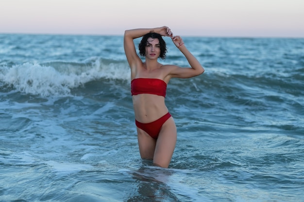 Summertime recreation concept. Beautiful young sexy woman with fit trained slim body wearing red swimwear bikini poses on a sandy beach. Fashion female model near the sea.