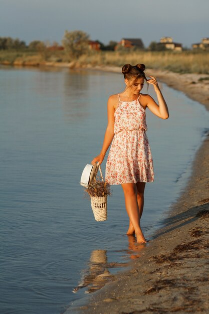 the sun is shining on the model. girl at sunset by the sea with a straw bag and a hat goes barefoot. The background is blurred.