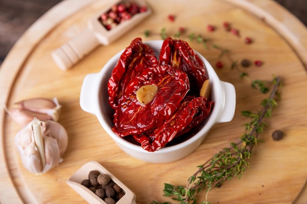 Sundried tomatoes in olive oil in a white bowl with pepper garlic and thyme on a wooden board Home cooking rustic style Selective focus closeup