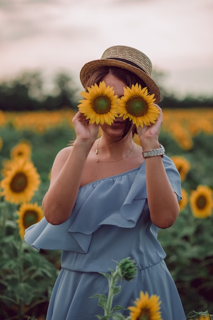 Sunflower eyes. Dreaming young woman in blue dress and hat holding sunflowers like eyes in a field of sunflowers at summer, front view. Looking forward. copy space