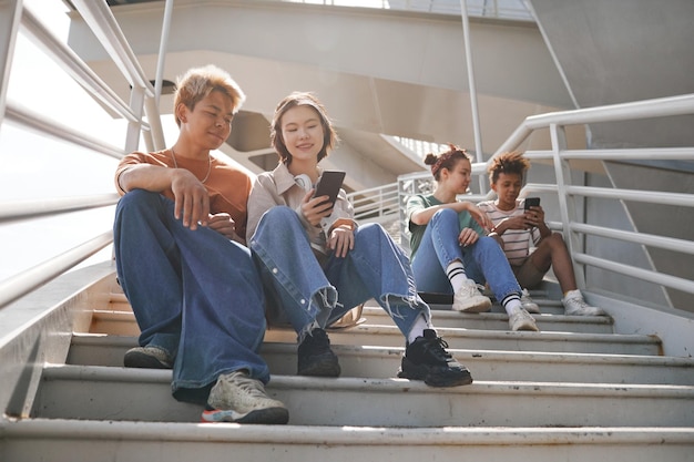 Photo sunlit full length shot of diverse teenagers using smartphones outdoors while sitting on metal stairs