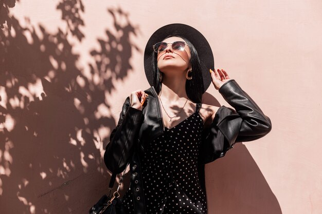 Sunny portrait pretty young brunette woman in elegant hat in beautiful dress in fashionable sunglasses in leather black jacket with dress near pink wall on beach. Sexy girl in trendy outfit