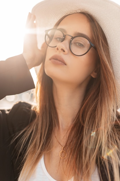 Sunny portrait of a pretty young fashionable woman in glasses and a white hat with a black jacket and t-shirt at sunset. Beachwear Women's Clothing Collection